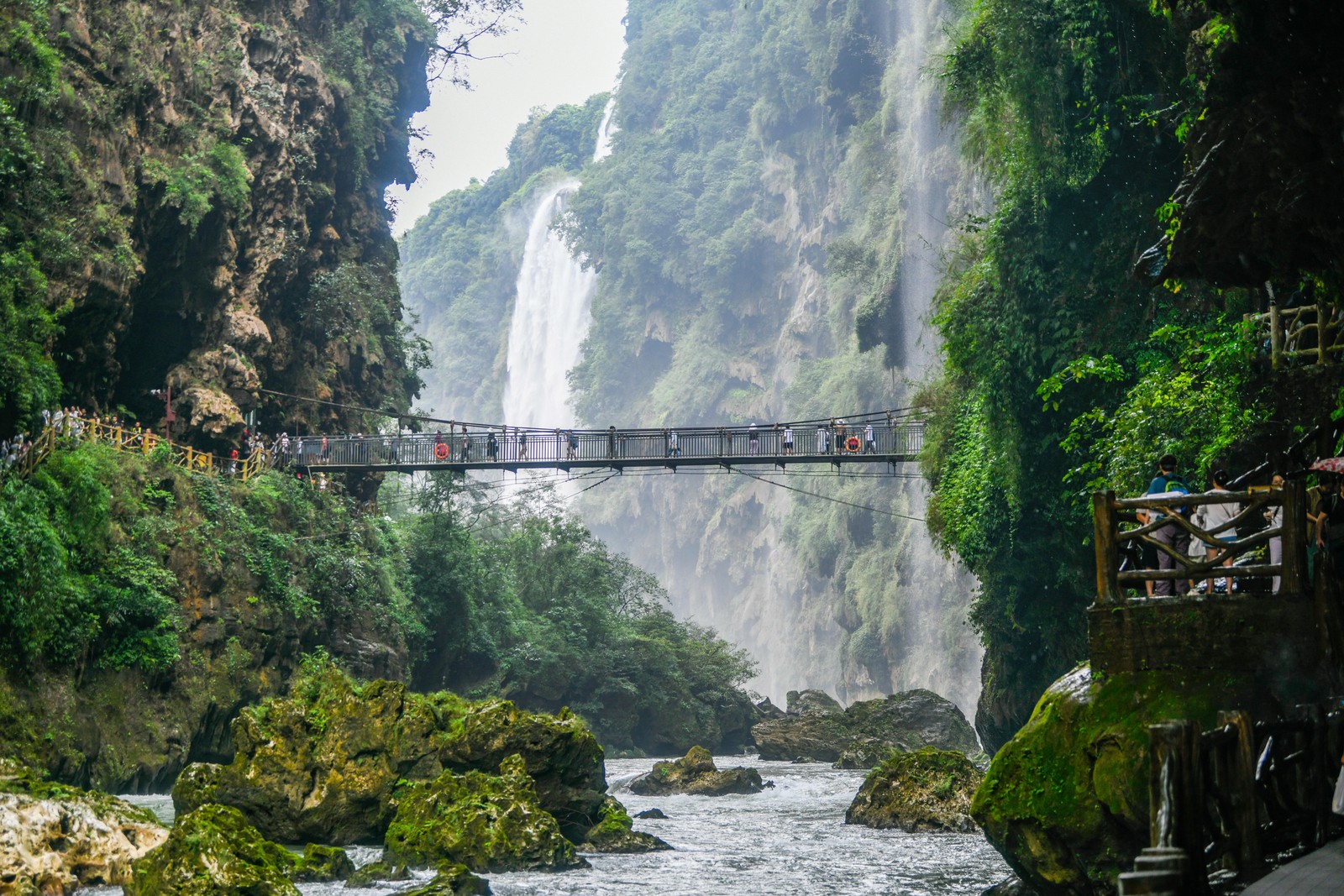 Tourists walk on a footbridge suspended between two tall cliff walls.