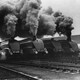 Five black steam engines blow smoke into the air.