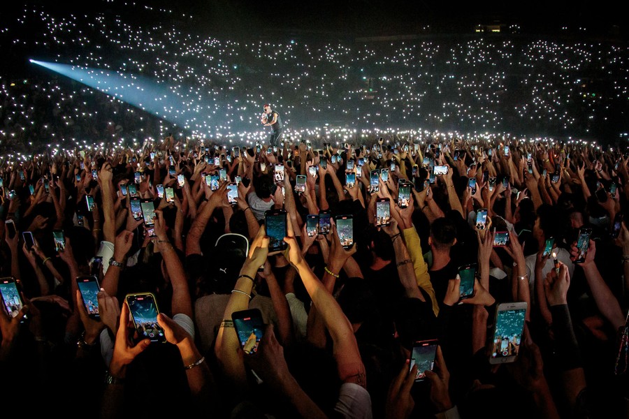 Hundreds of audience members in a stadium hold up phones during a performance.