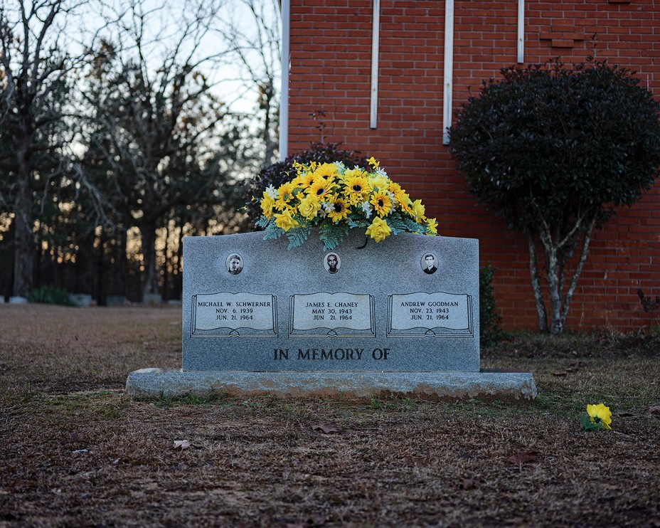 photo of memorial stone with "In Memory Of" and photographs and names of Schwerner, Chaney, and Goodman, with yellow flowers on top and brick church in background