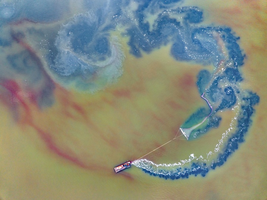 A drone's-eye view of a small boat towing fishing apparatus through colorful salt water, stirring up the water.