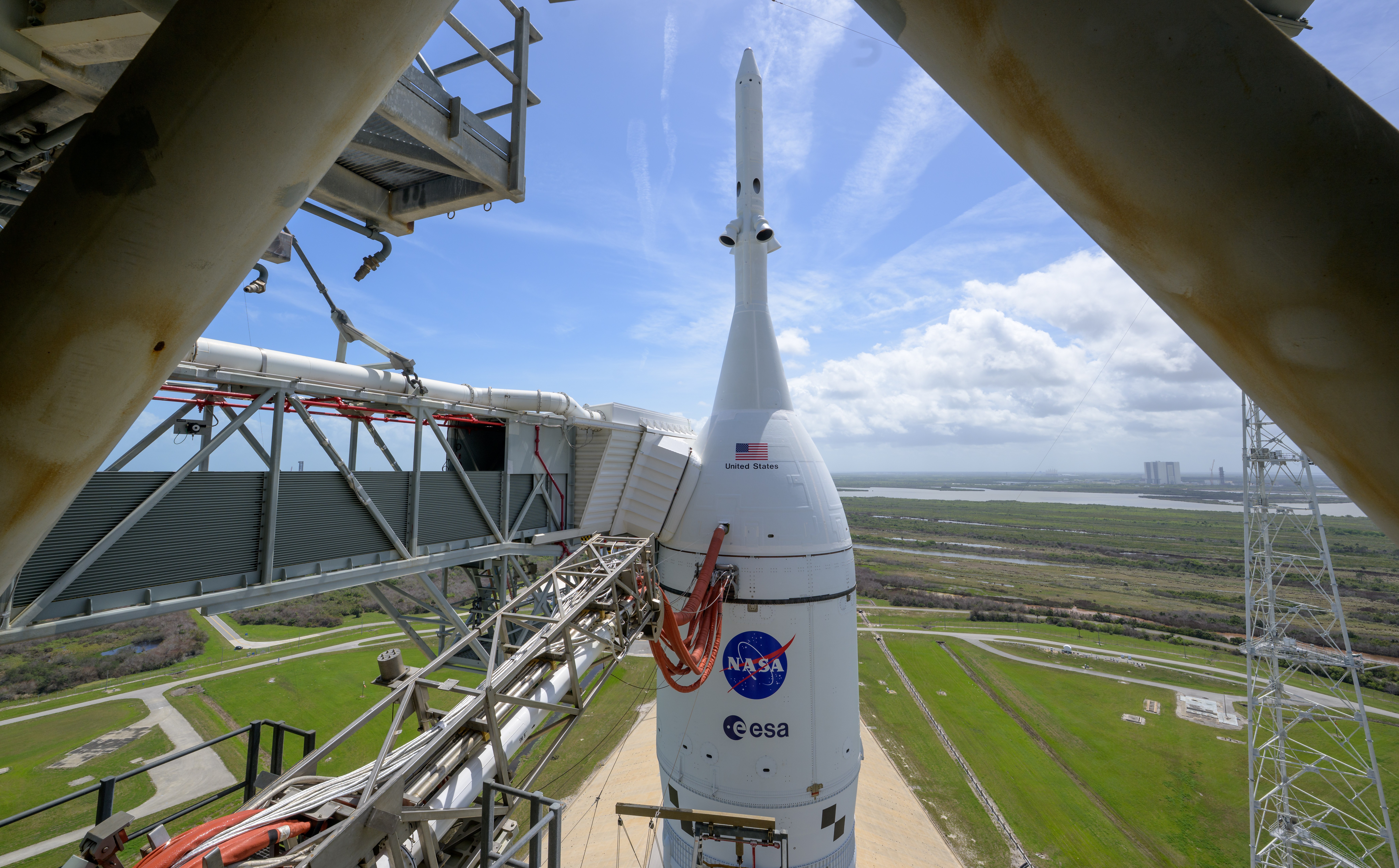 A view of the crew module atop a tall rocket, seen from a tall support structure beside the rocket.