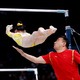 A gymnast falls face-down from a high bar she had been swinging from as a coach reaches out one arm to slow her.