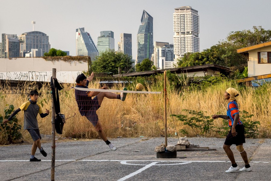Three people are seen kicking a ball over a net in an outdoor playground.