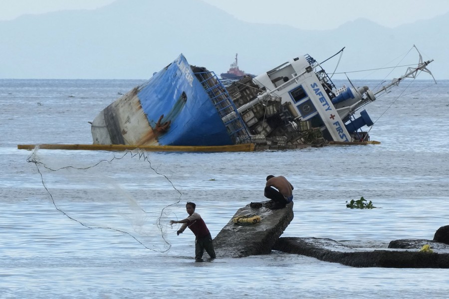 A fisherman throws a net near a listing cargo ship.
