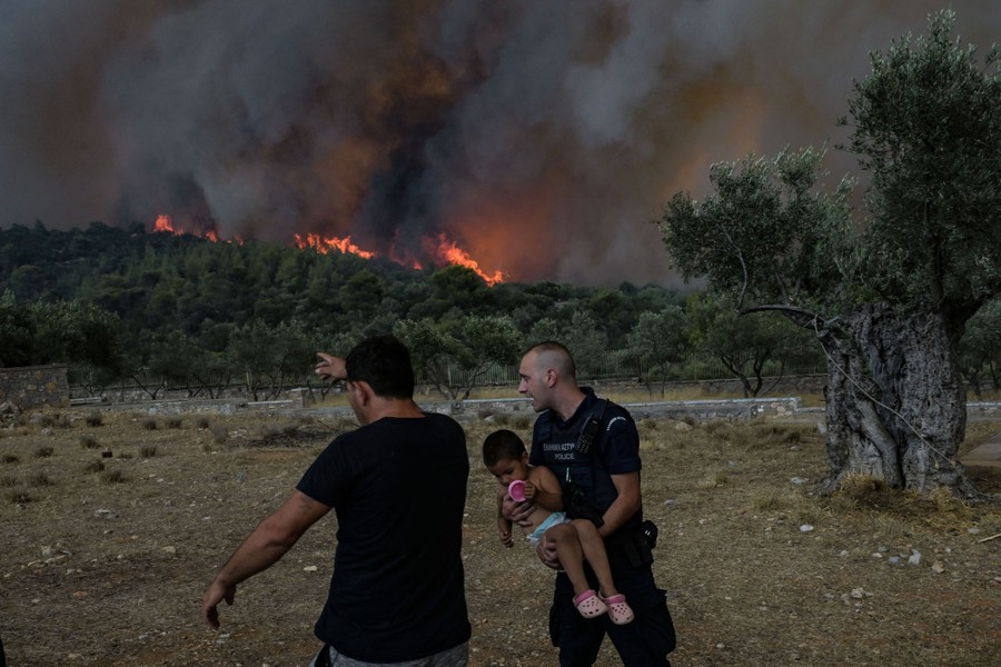 A police officer carries a child away, as a large wildfire burns on a wooded hill in the background.