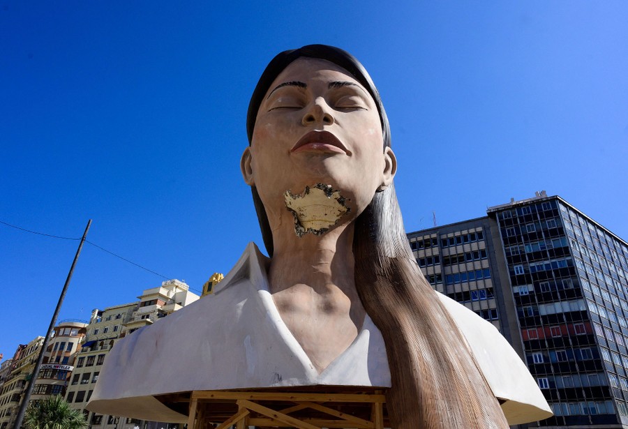 A damaged large structure of a woman stands in front of buildings.