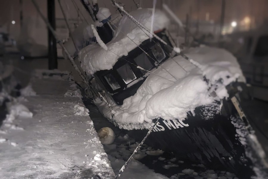 A small boat, covered in heavy snow, leans over dangerously while moored in a harbor.