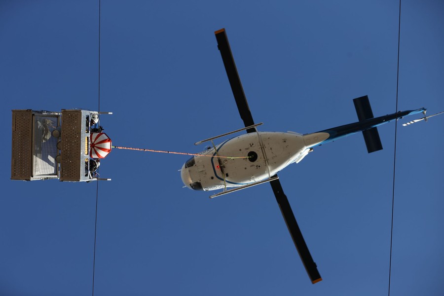 A view from directly below a helicopter that dangles a basket carrying workers who are installing a marker ball on a power line.