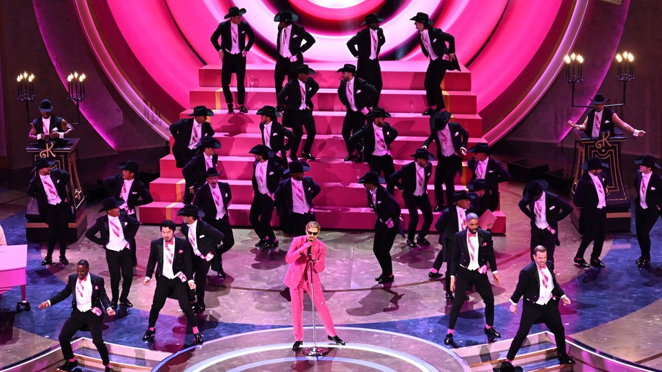 Ryan Gosling, dressed as Ken in a pink suit and gloves, sings into a microphone on the Oscars stage as several dozen men in black tuxedos dance behind him.