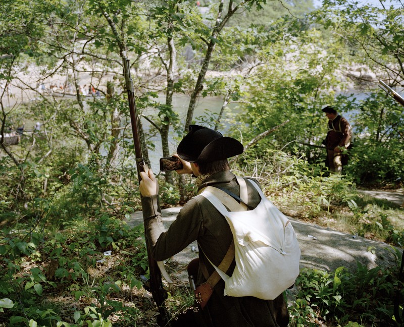 photo of a young reenactor of an American soldier filming the English soldiers arriving with his iPhone from an overlook while holding his musket in his left hand, with another American reenactor in the background
