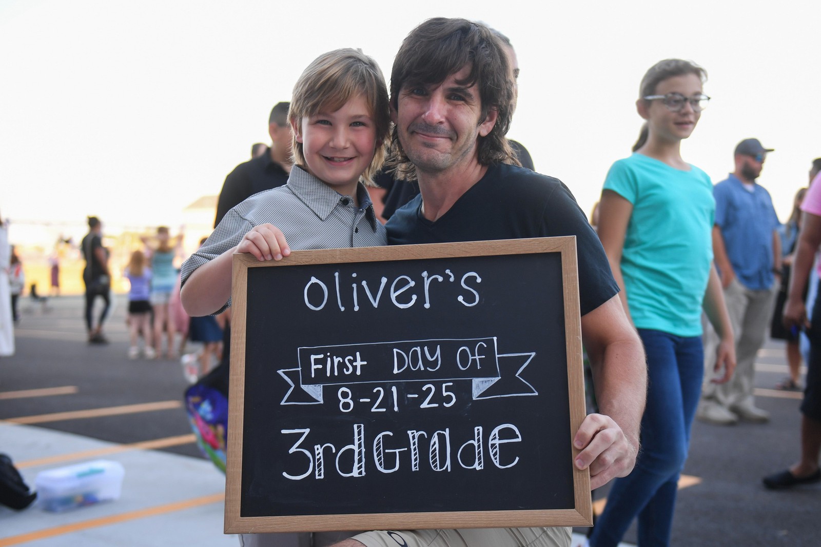 A father and son pose outside of a school, holding a small chalkboard that reads 