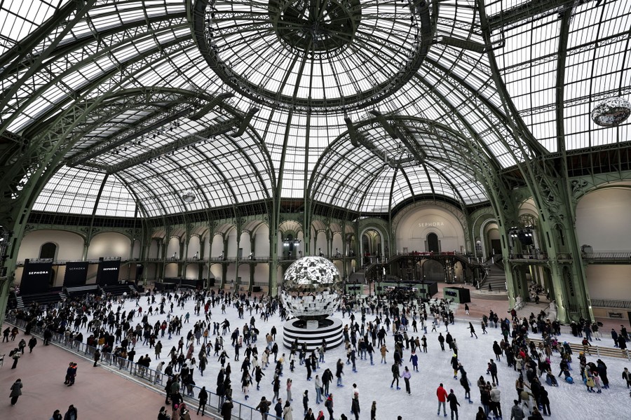 People skate on an enormous indoor ice rink beneath arched iron supports holding up hundreds of glass roof panels.