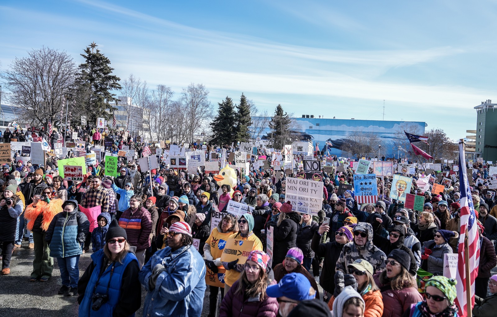 A group of protesters wearing warm clothing stand together in a city square holding signs