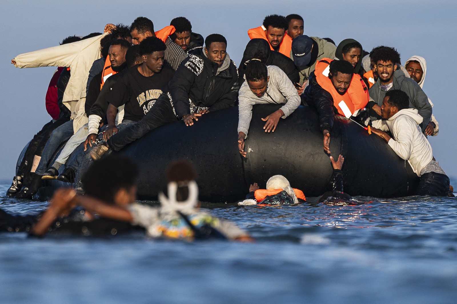 People in an overcrowded inflatable boat reach down to try and help several swimmers to climb aboard.