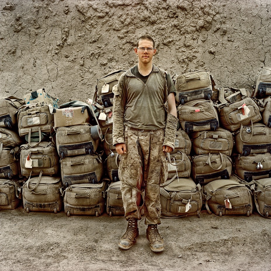 Soldier with torn shirt and pants standing in front of a pile of khaki-colored wheeled duffel bags