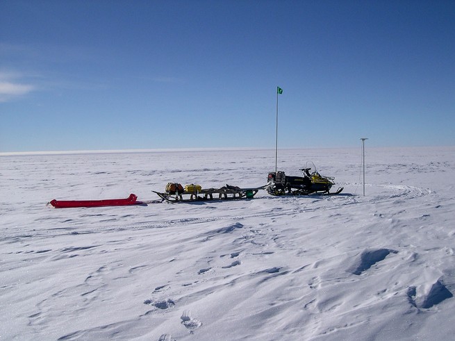 photo of snow field with snowmobile trailing sleds and pole sticking up from ground