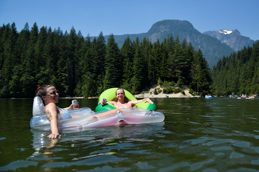 People float in inflatable chairs, smiling and enjoying beverages, surrounded by pine trees and mountains.