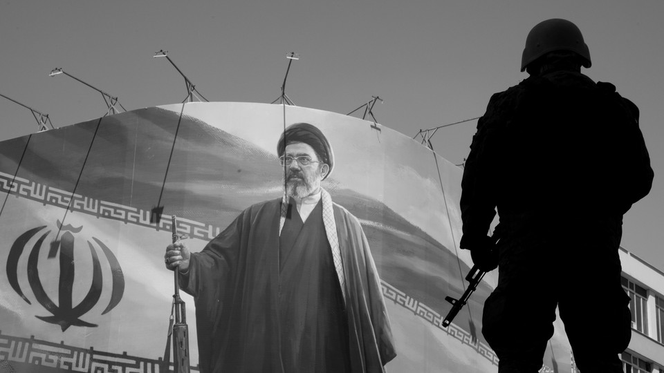 Black-and-white photograph of an armed soldier standing in front of a mural of Mojtaba Khamenei holding a rifle next to the Iranian flag