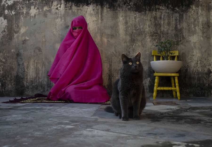 A woman in a colorful hijab sits against a wall beside a cat.