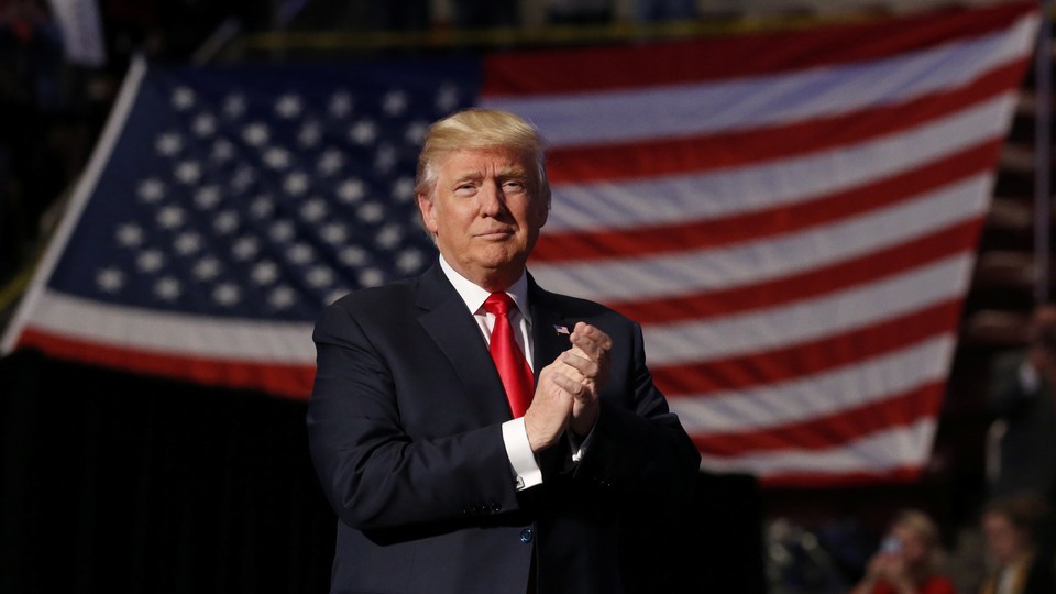 President Donald Trump stands in front of an American flag with clasped hands.