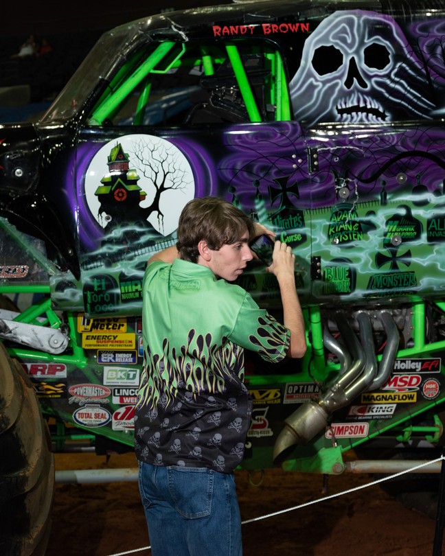 A young man stands beside a monster truck that features a skull on its side.