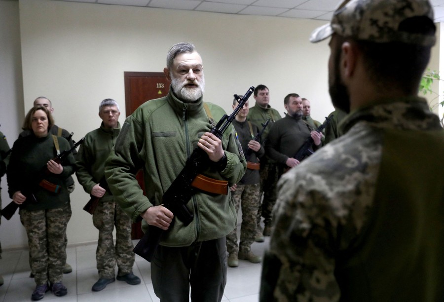 About a dozen people stand at attention in a room, holding rifles, in front of a soldier.