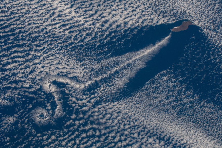 An island leaves a turbulent wake as the wind pushes clouds past, seen from orbit