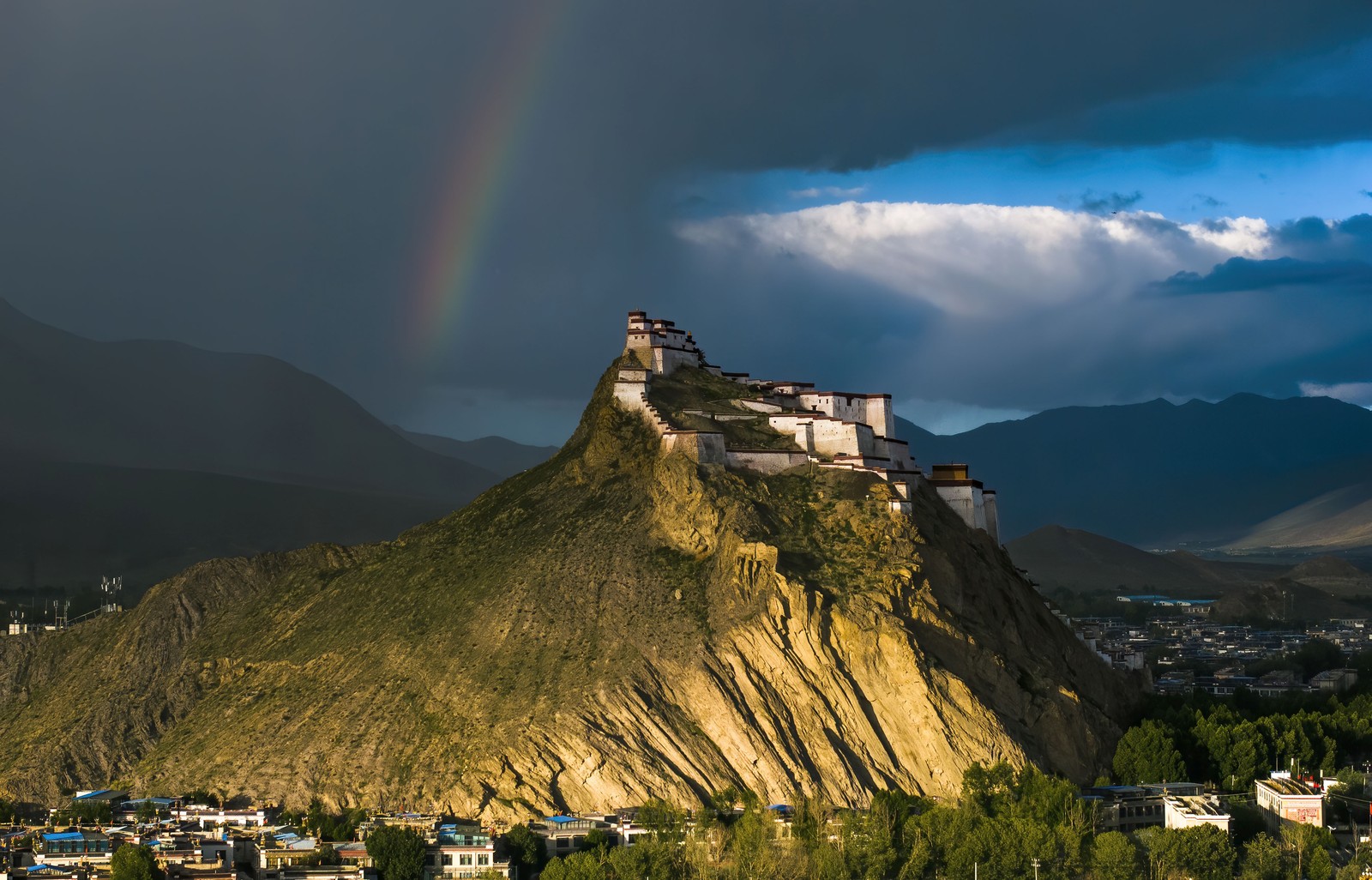 A rainbow appears in a dark sky above a historic religious structure on a rocky hilltop.