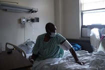 A masked man in a green gown sits up in a hospital bed, wearing medical cuff around his upper arm