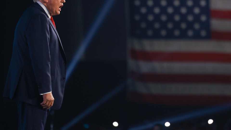 Color photograph of Donald Trump speaking onstage, with his head cut off mid-nose.