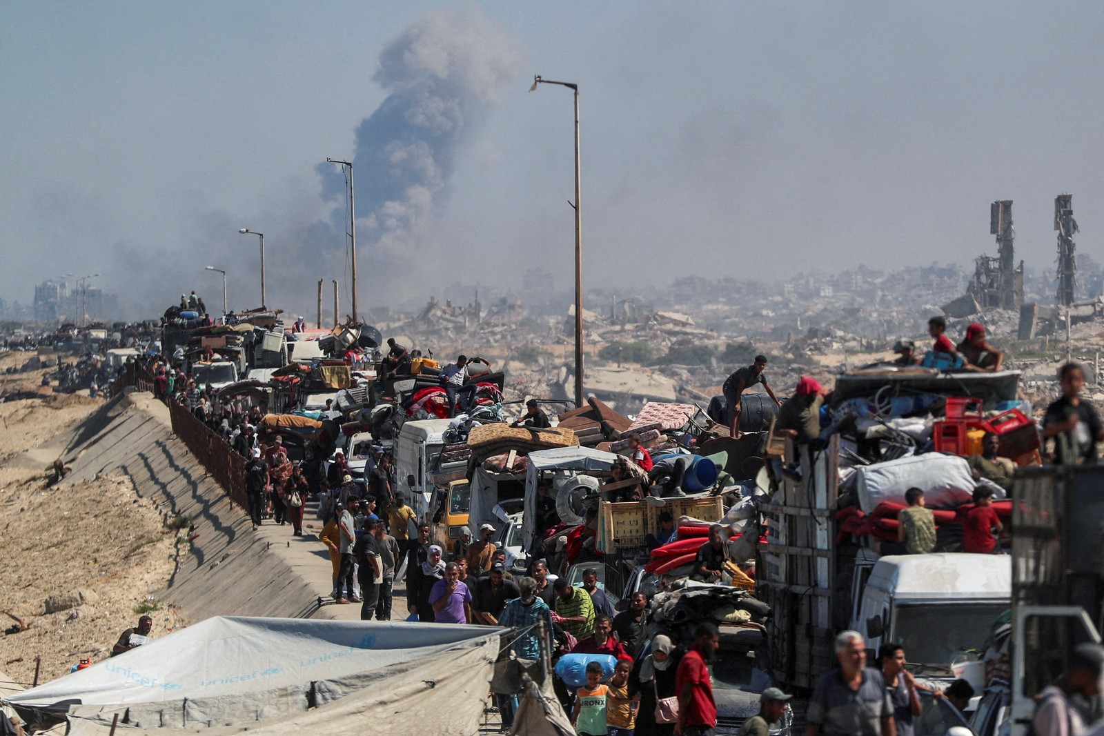 Hundreds of Palestinians ride on and walk near a very long line of packed trucks and cars on a road leading away from ruined buildings and smoke rising in the distance.