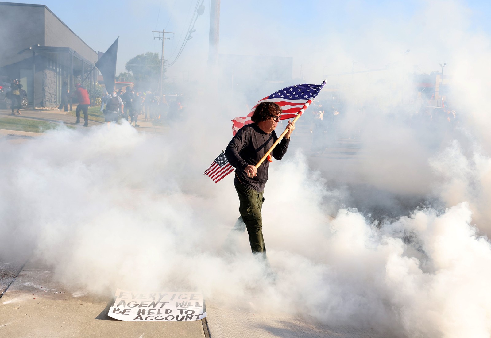A person carries an American flag as they walk through a cloud of tear gas at a protest outside of an ICE facility.