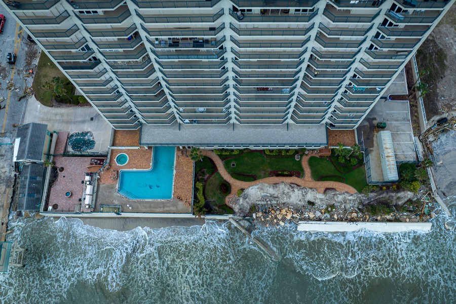 An aerial view of waves splashing against a damaged seawall beside a tall building.