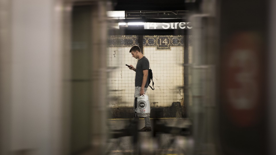 Seen between the cars of a passing train, a man looks at his phone on a subway platform.