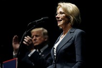 Betsy DeVos stands at a lectern in the foreground of the photo. Donald Trump is clapping behind her.