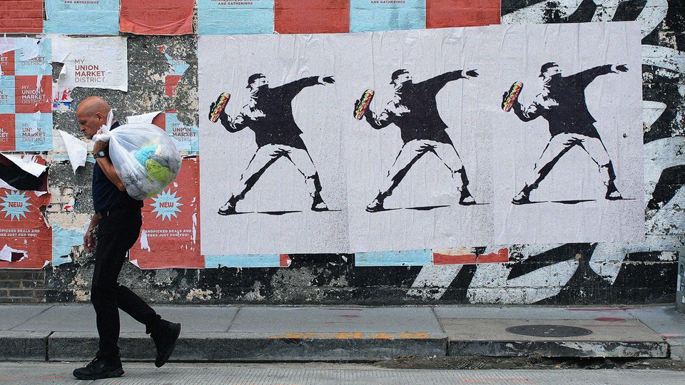 A city wall displays three copies of an image rendering the Sandwich Thrower in the style of Banksy's "Flower Thrower," while a man with a plastic bag walks in the foreground.