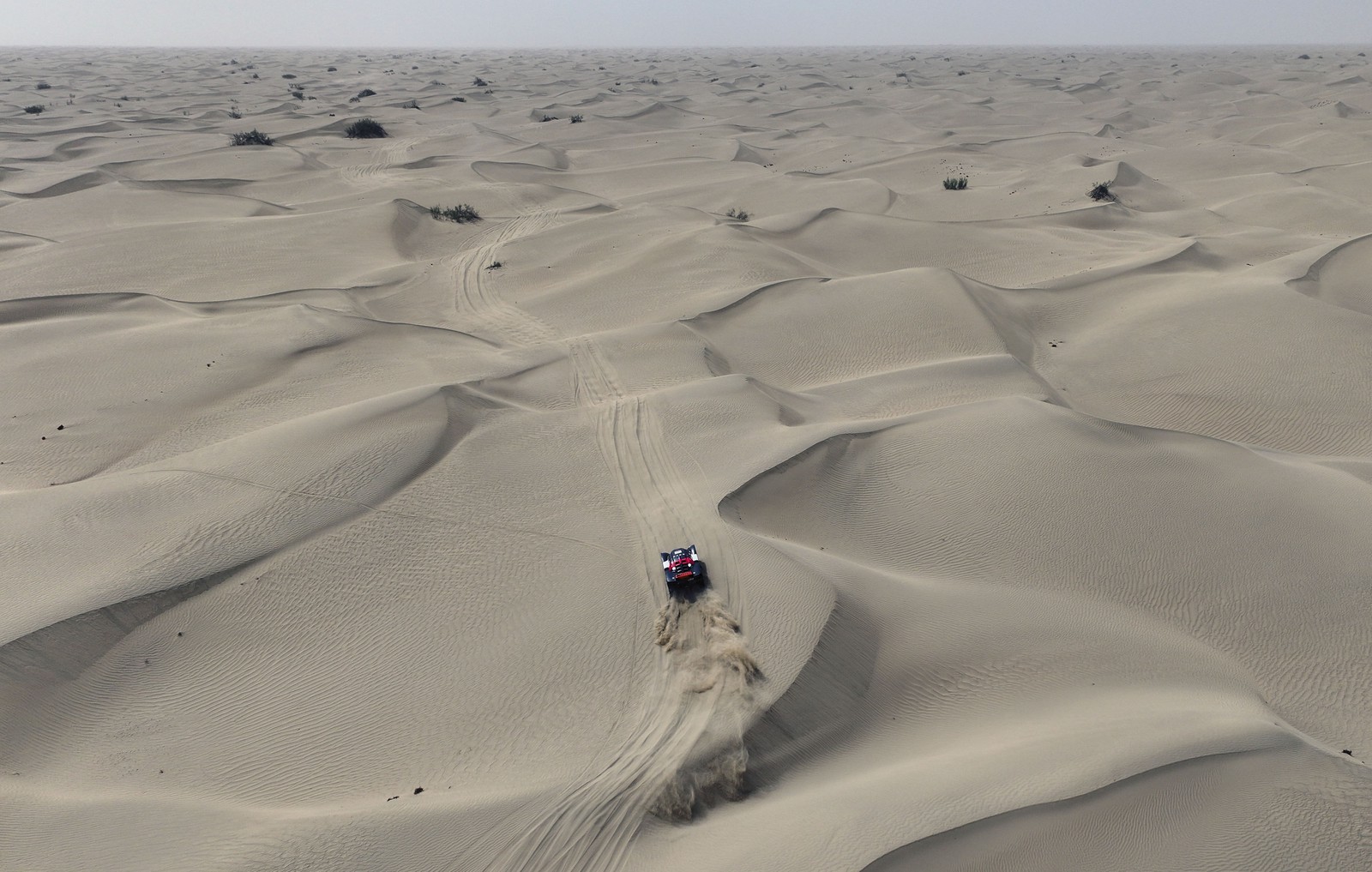 An aerial view of a rally race car driving across a broad field of sand dunes