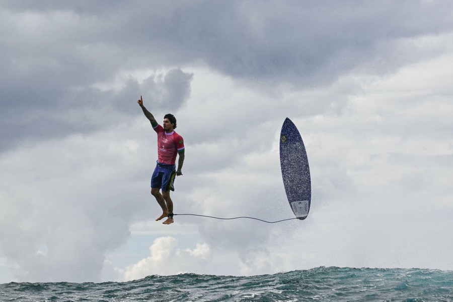 A surfer points upward while jumping above a wave with his surfboard beside him, appearing to stand casually upright in midair.