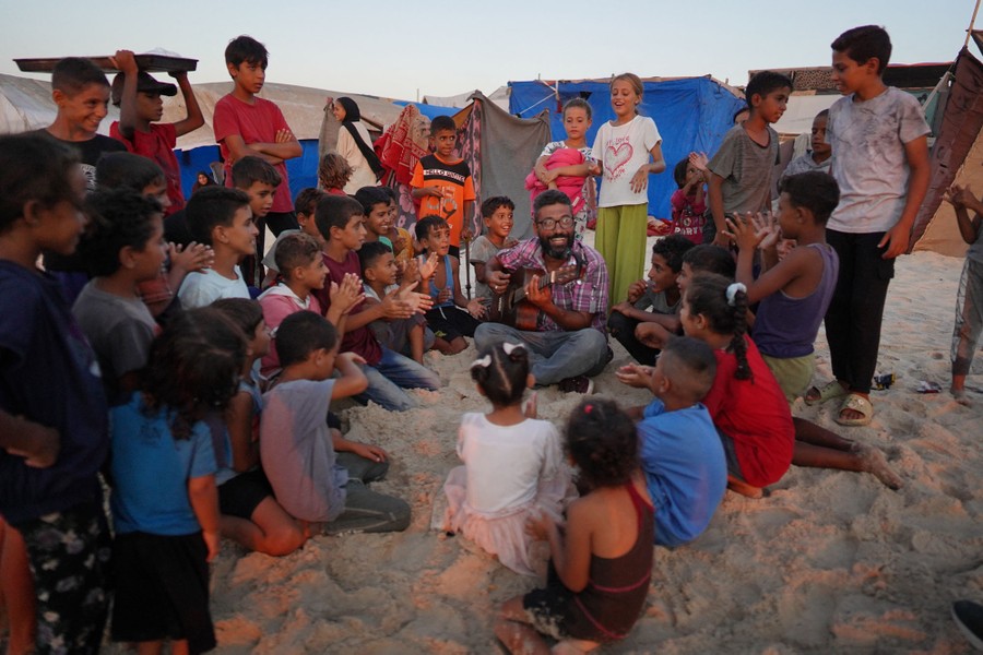A music teacher sits on a beach, playing guitar, surrounded by many children who smile and clap.