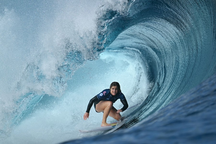A surfer kneels low on her board, riding through the curl of a large crashing wave.