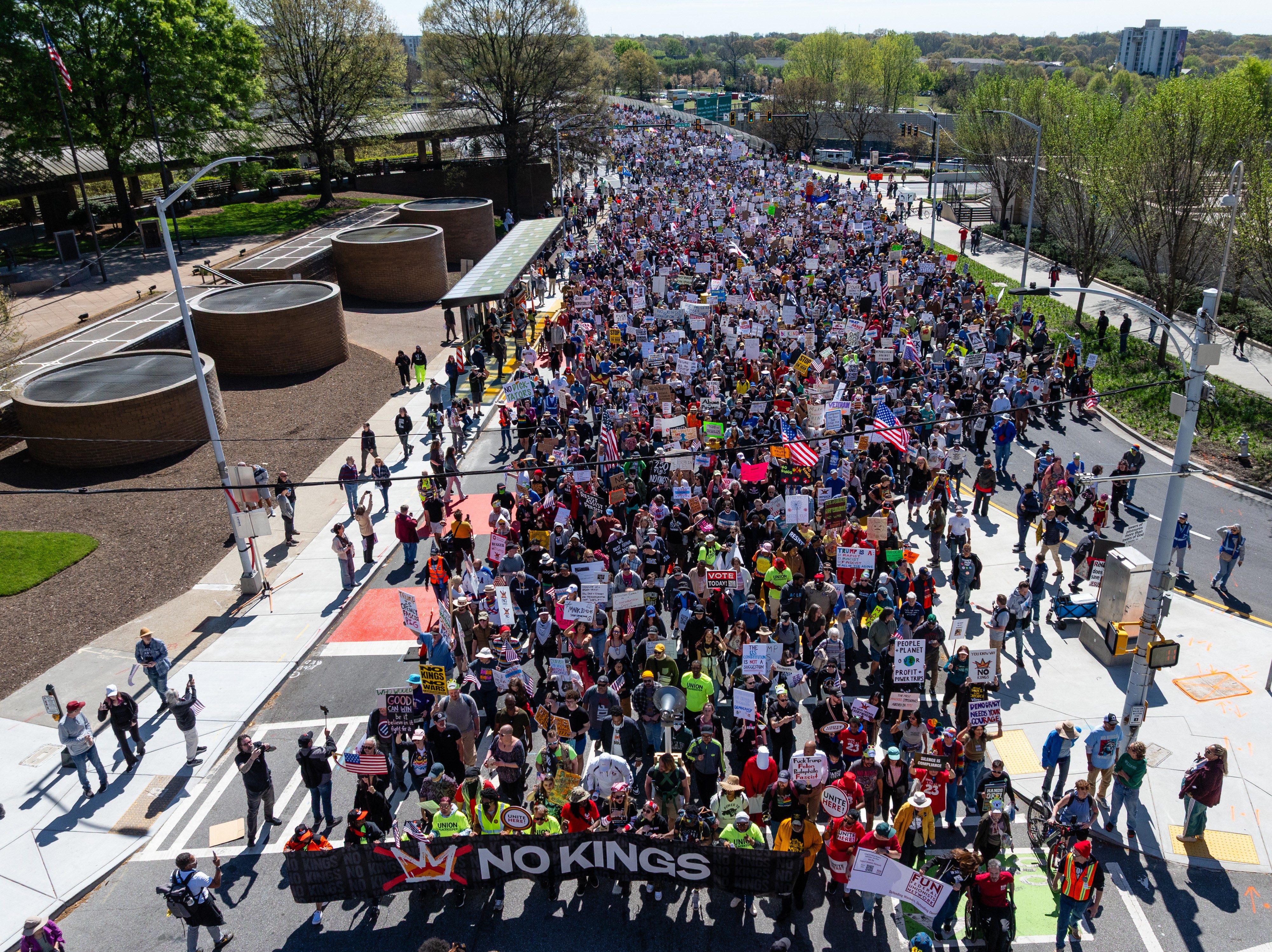 A group of thousands of protesters march down a city street, behind a banner carried at front, which reads