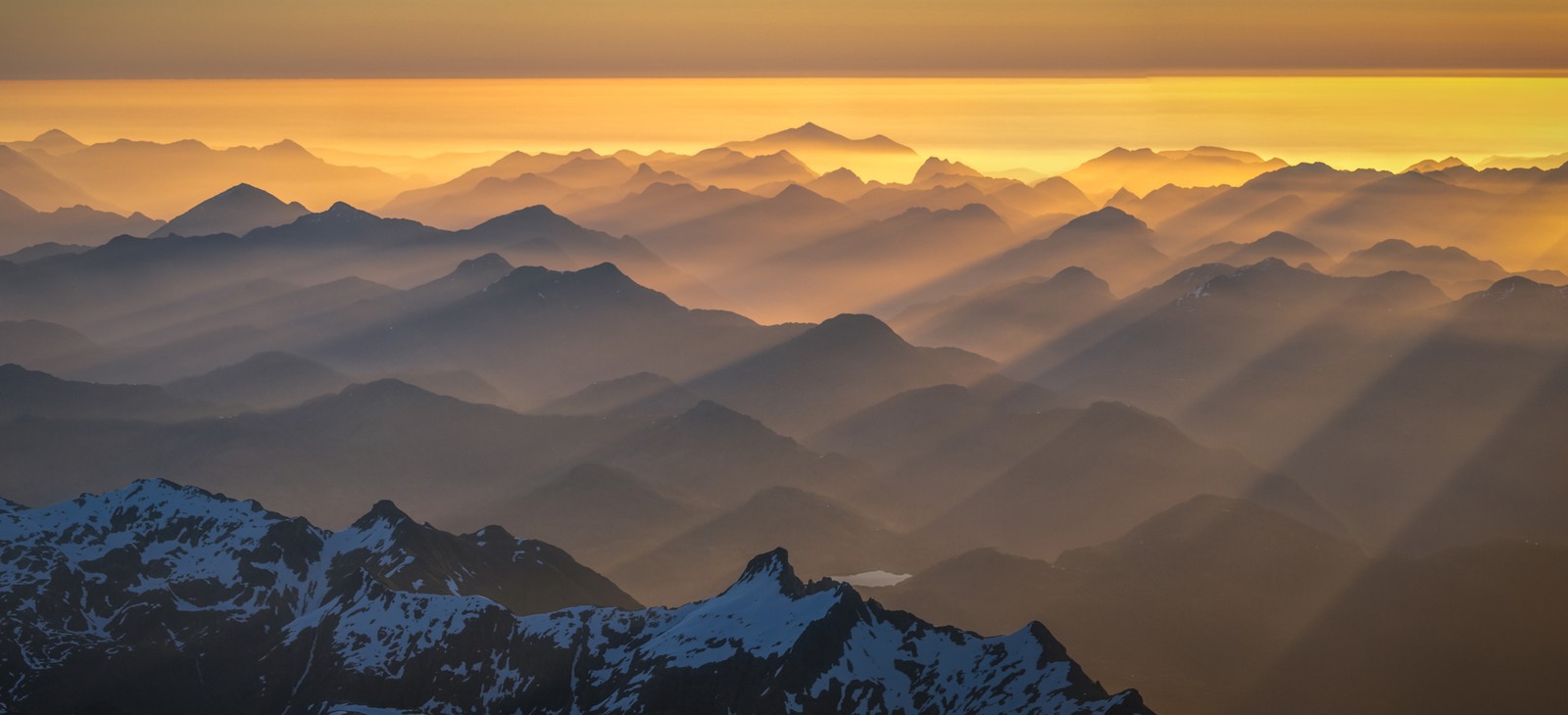 Rays of sunlight filter through fog and valleys among many mountain peaks, seen from above.