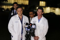 Two doctors in white coats stand in front of microphones