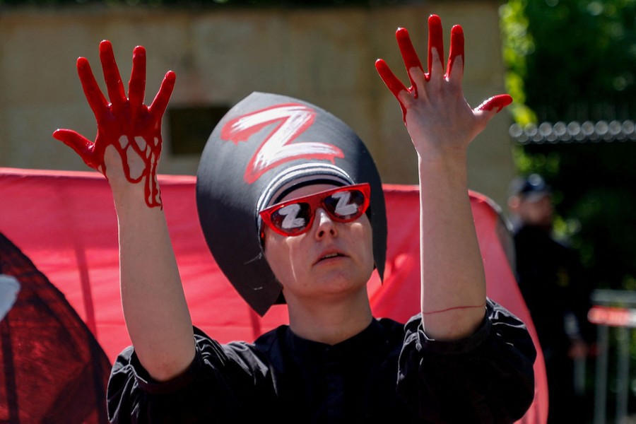 An activist stages a protest, wearing a large hat with a "Z" on it, and with blood dripping from their hands.