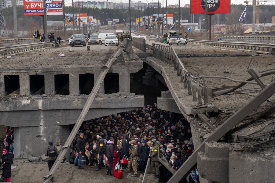 A large crowd of people stands together under a destroyed bridge as they try to flee a war zone.