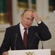 Vladimir Putin stands at a lectern, looking downward and holding his hand up to his head.