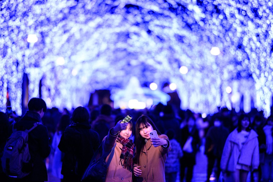 Two people take selfies in front of an avenue of illuminated trees.