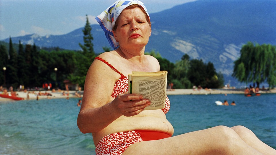 A woman in a kerchief and red bikini reads a book on a beach.