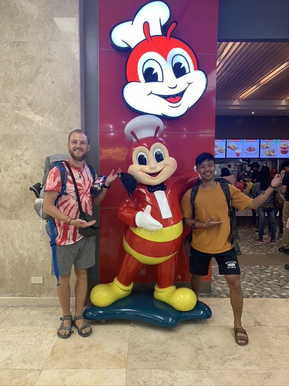 Two men stand outside a fast food restaurant, next to a statue of a smiling bug wearing a chef's hat and a red and yellow outfit. The men are mimicking the statue's pose.